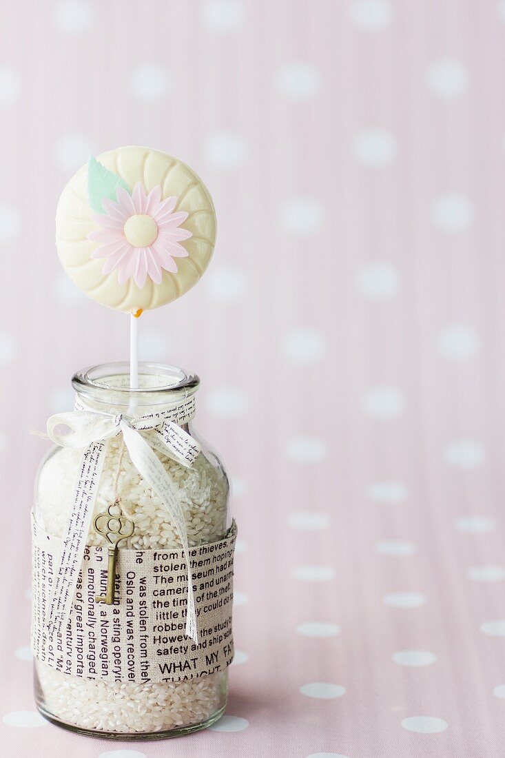 A carrot lolly decorated with a sugar flower in a jar of rice