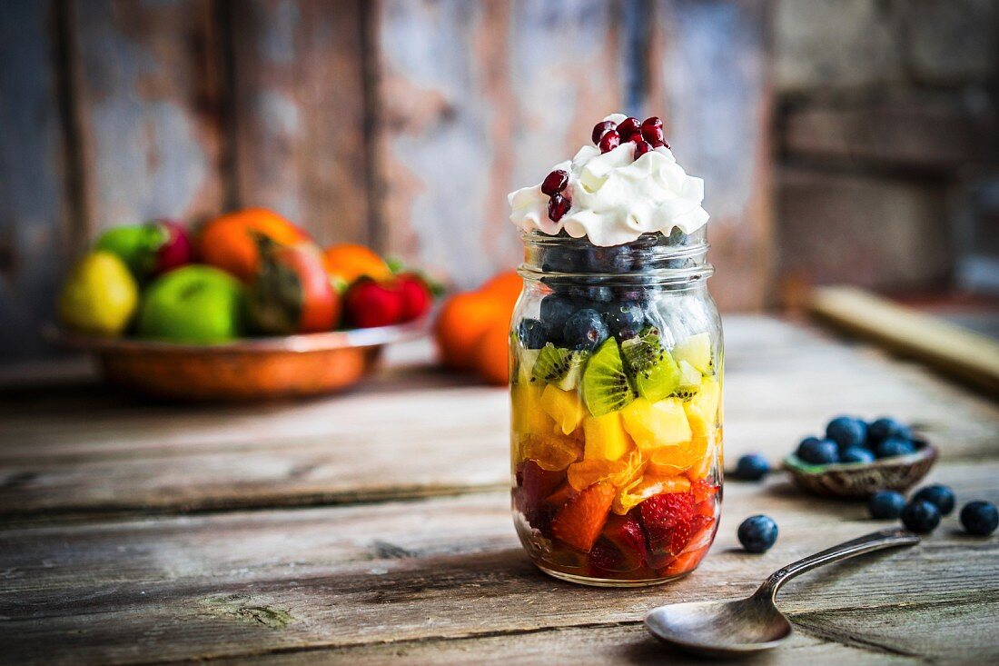 A colourful fruit salad in a jar topped with cream and pomegranate seeds on a rustic wooden surface