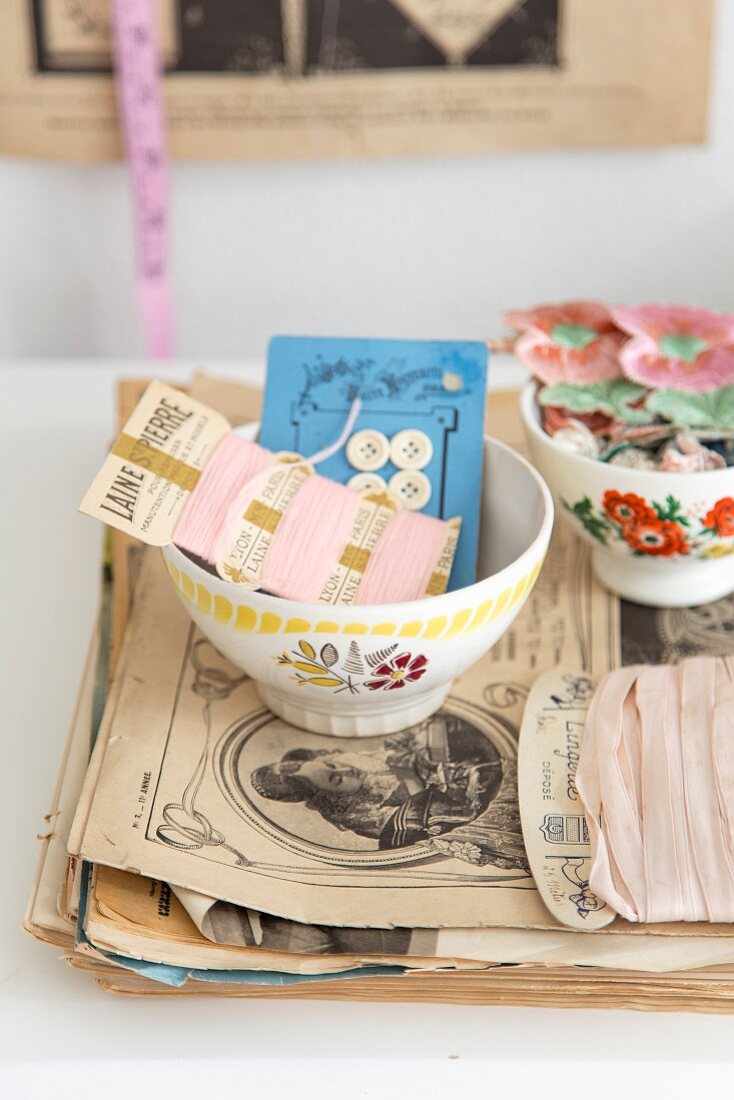 Still-life arrangement of French bowls and vintage sewing utensils on stack of antique magazines