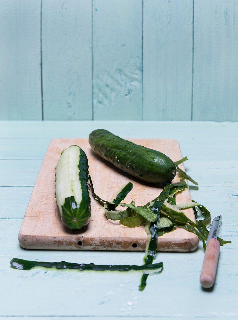 A peeled cucumber on a wooden board with a peeler and peel