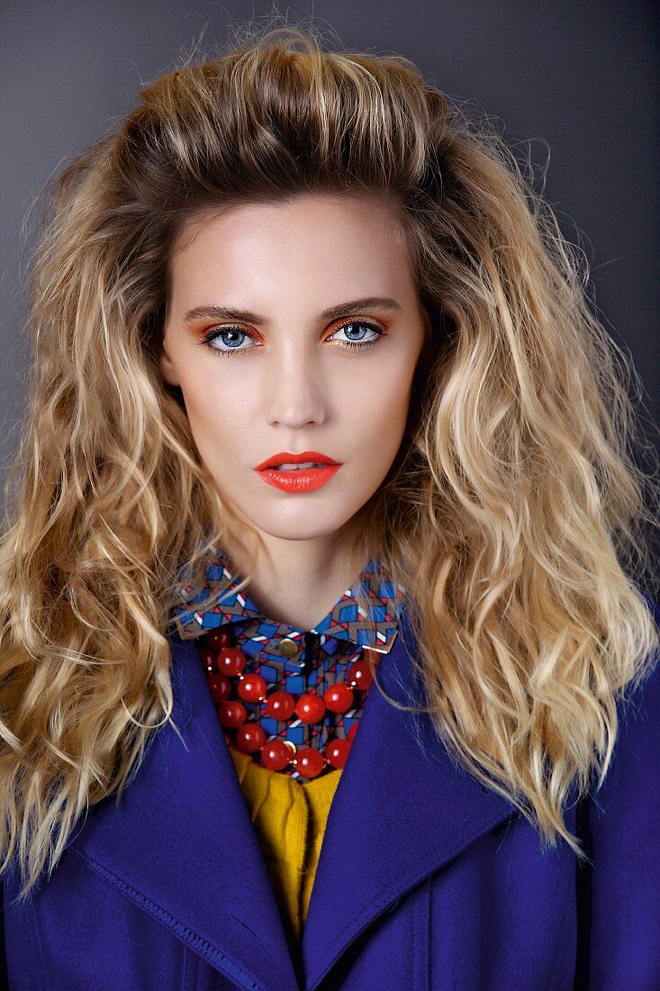 A blonde woman wearing a blouse, a jumper, a jacket and the necklace in various colours