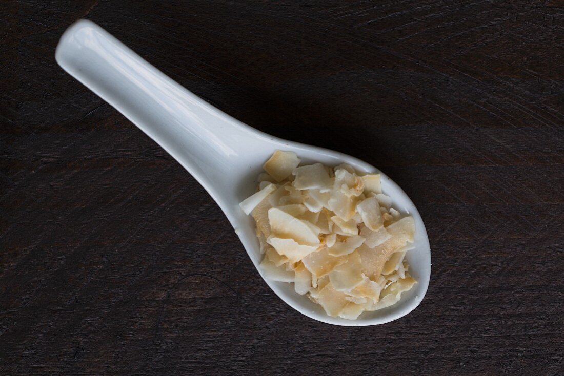 Coconut chips on a porcelain spoon on a dark wooden surface