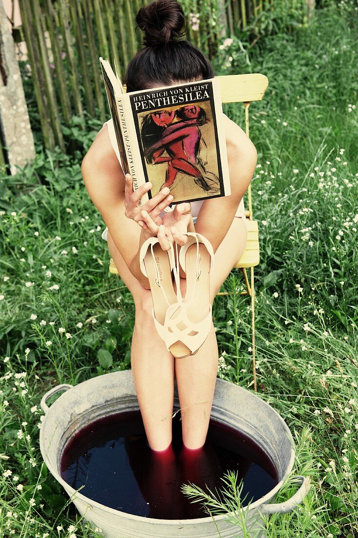 A woman taking the foot bath and holding a book and her sandals in her hands