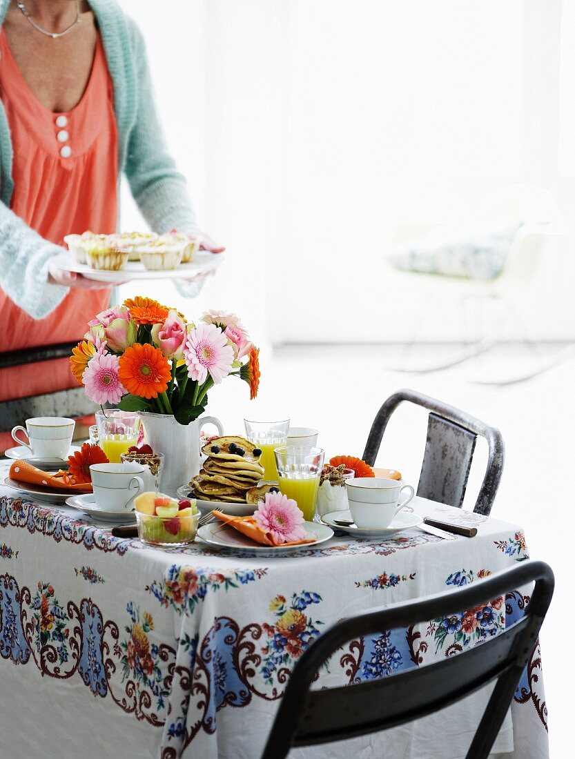 A table laid for breakfast with a bunch of colourful gerberas and a woman holding a plate of muffins