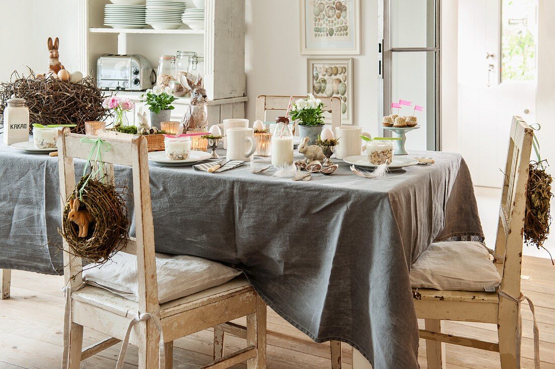 Table set for Easter breakfast with grey tablecloth and decorative wreaths on backrests of vintage chairs