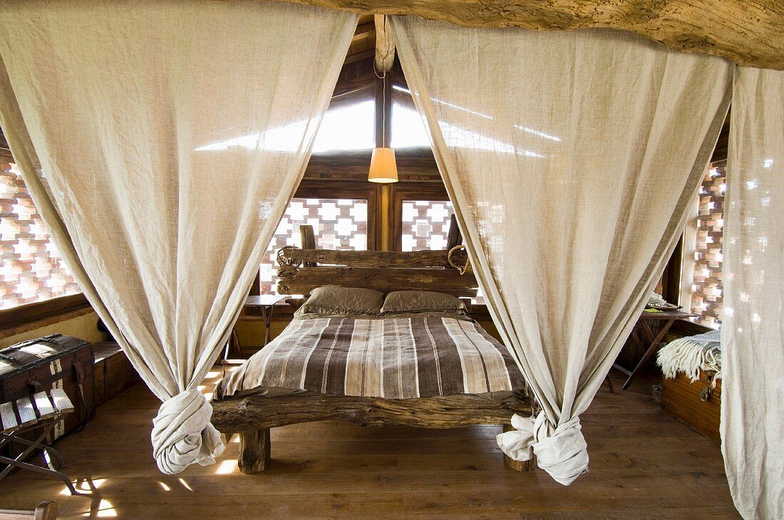 Bed frame made from plain wooden beams with linen curtains knotted at lower ends in rustic bedroom with