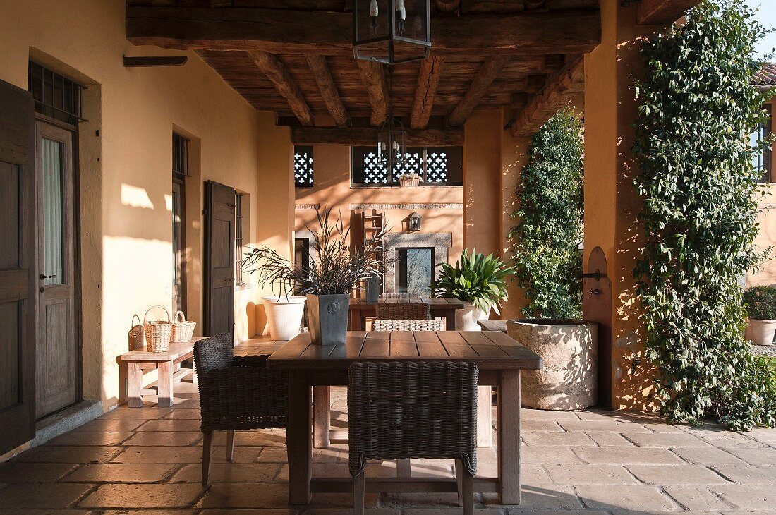 Wicker chairs around simple wooden table on roofed terrace adjoining house with walls painted apricot