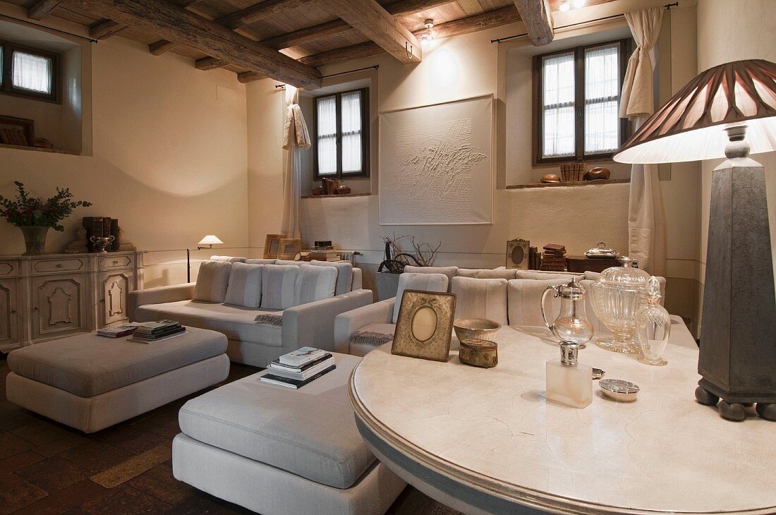 Table lamp on round table, elegant white sofa set and matching ottomans in renovated rustic living room