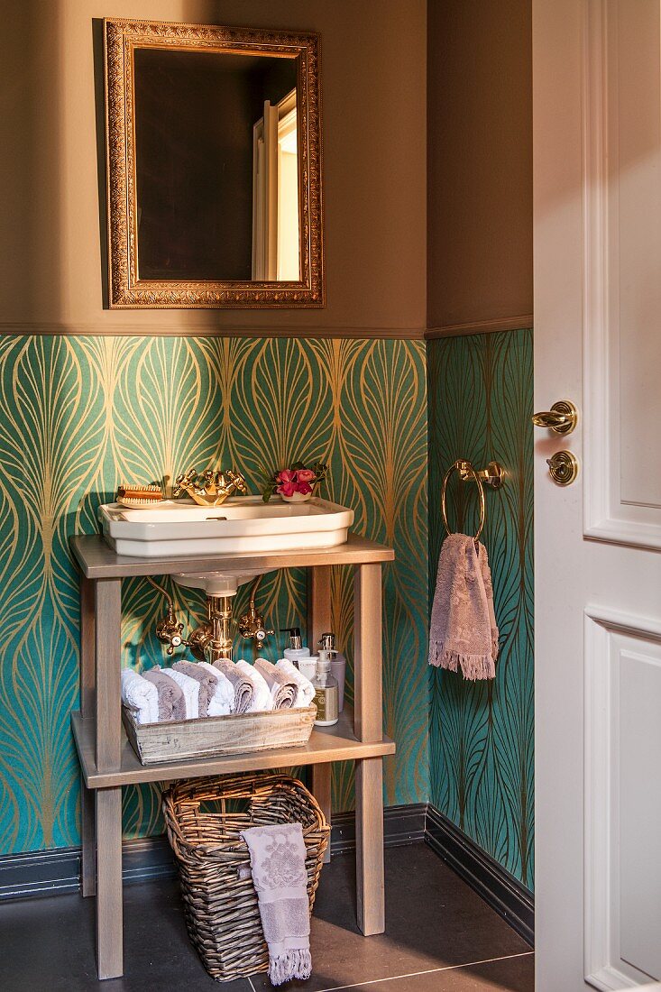 Sink on wooden frame against dado with turquoise and gold patterned wallpaper below gilt-framed mirror on brown-painted wall