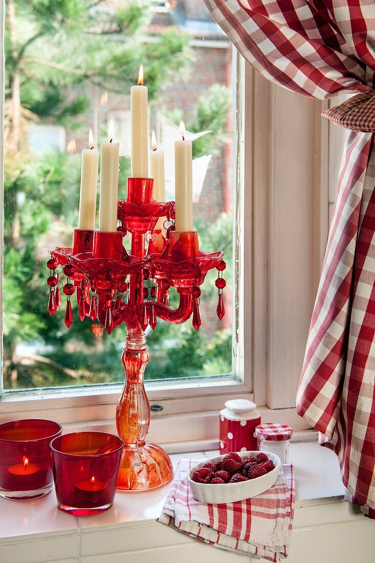 Roter Glas-Kerzenleuchter und Teelichtgläser auf Fensterbrett mit rot kariertem Vorhang