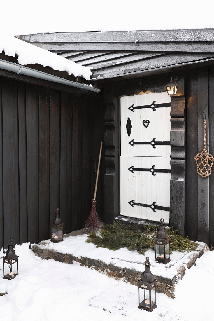 Lanterns in snow leading to black wooden hut with white front door