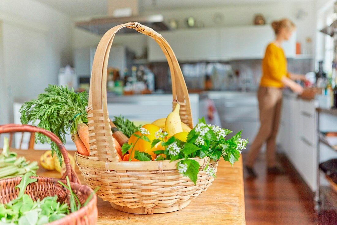 Baskets of fresh garden vegetables on a kitchen table with a woman in the background