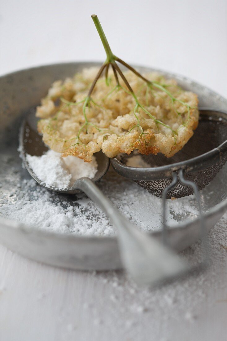Deep-fried elder flowers on a spoon and a tea strainer dusted with icing sugar
