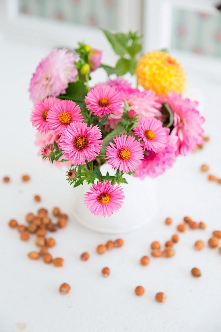Daisies and dahlias in white jug surrounded by scattered hazelnuts