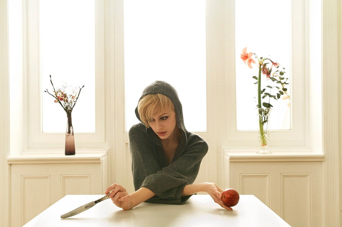 A young woman sitting on a table holding an apple and a silver knife