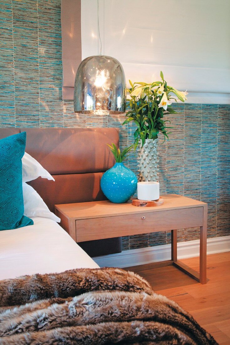 Shiny lampshade above bedside table, bed with light brown leather headboard and patterned wallpaper