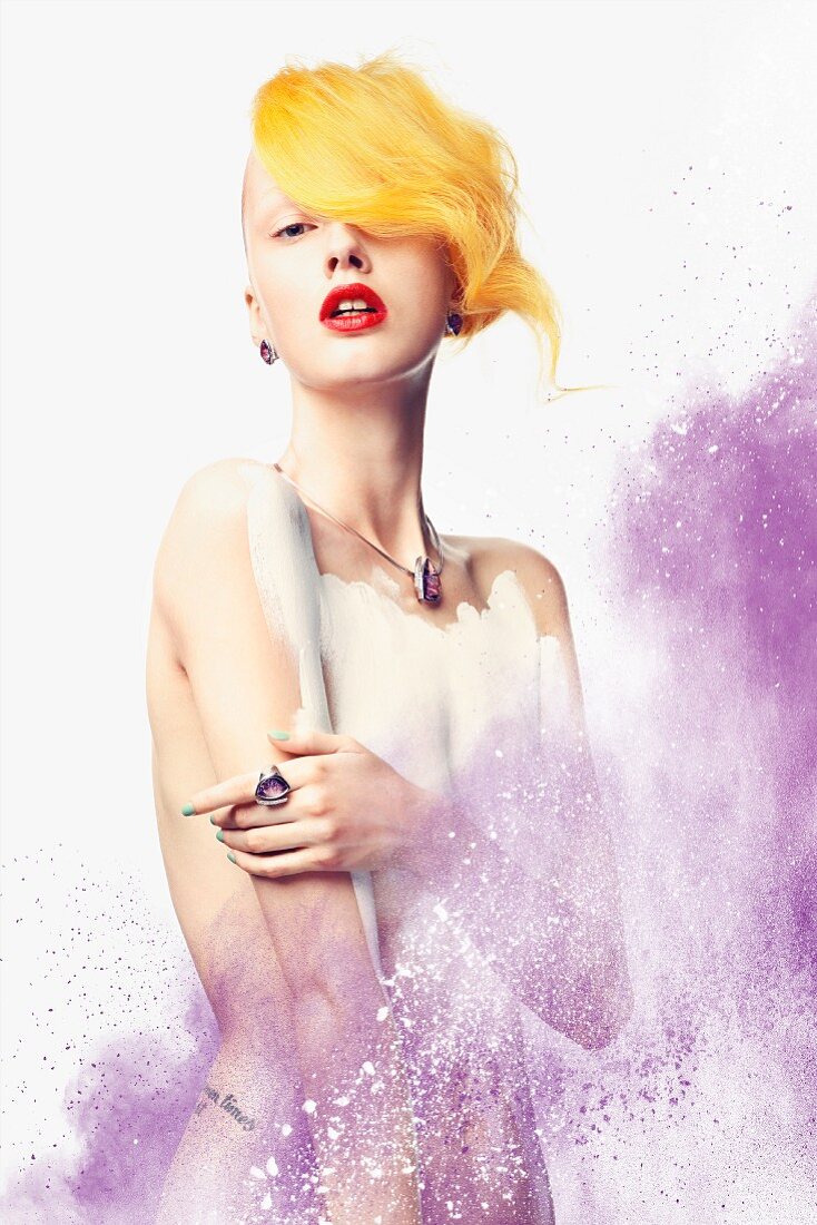 A young woman with a yellow hair piece, her body partially painted white, in a dusting of purple powder