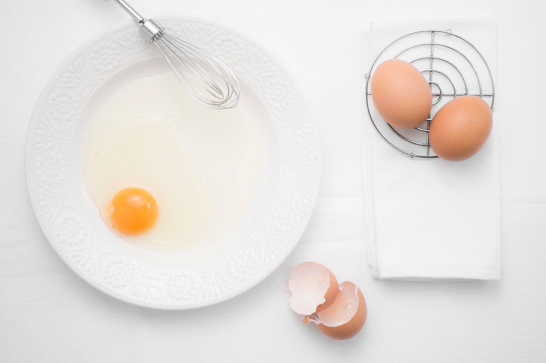 Aufgeschlagenes Ei mit Schneebesen auf Teller, daneben ganze Eier & Eierschalen