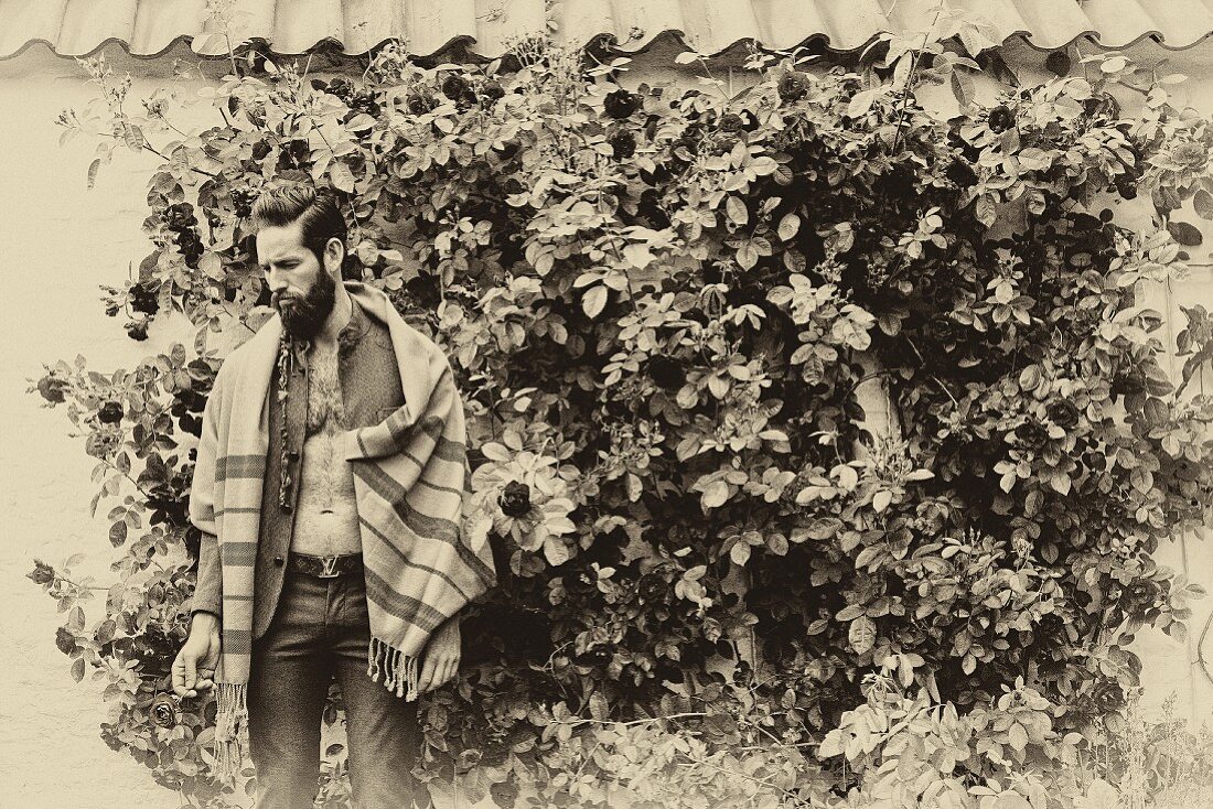 A man wearing a tweed jacket with a shawl over his shoulders standing in front of a rose bush