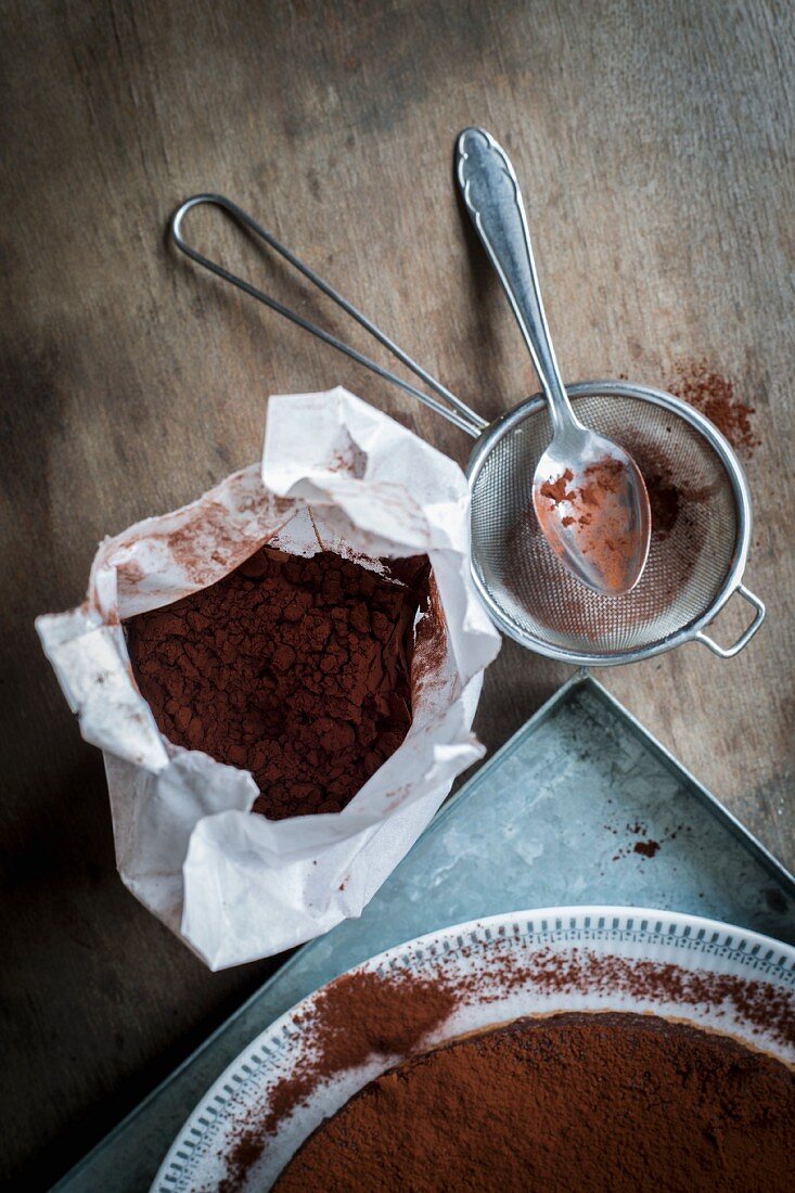 Chocolate cake on a metal tray with utensils for sprinkling cocoa powder on a wooden table