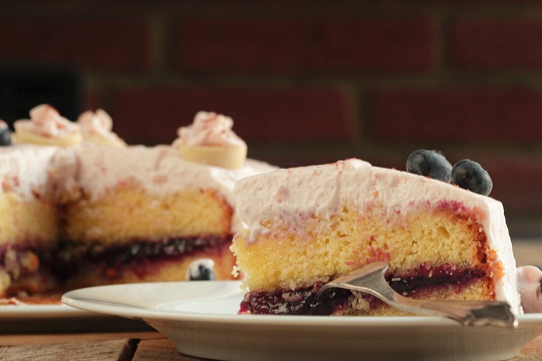 Ein Stück Heidelbeer-Sahne-Torte vor angeschnittener Torte