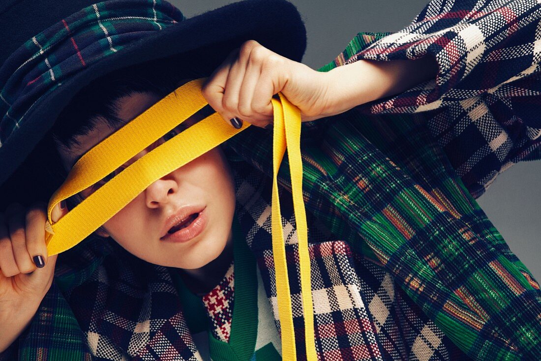 A young woman wearing an outfit made from various checked fabrics holding a yellow ribbon over her eyes