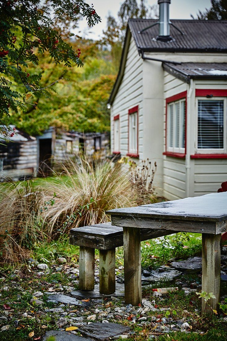 Rustikaler Tisch mit Bank in herbstlichem Garten, weisses Holzhaus im Hintergrund