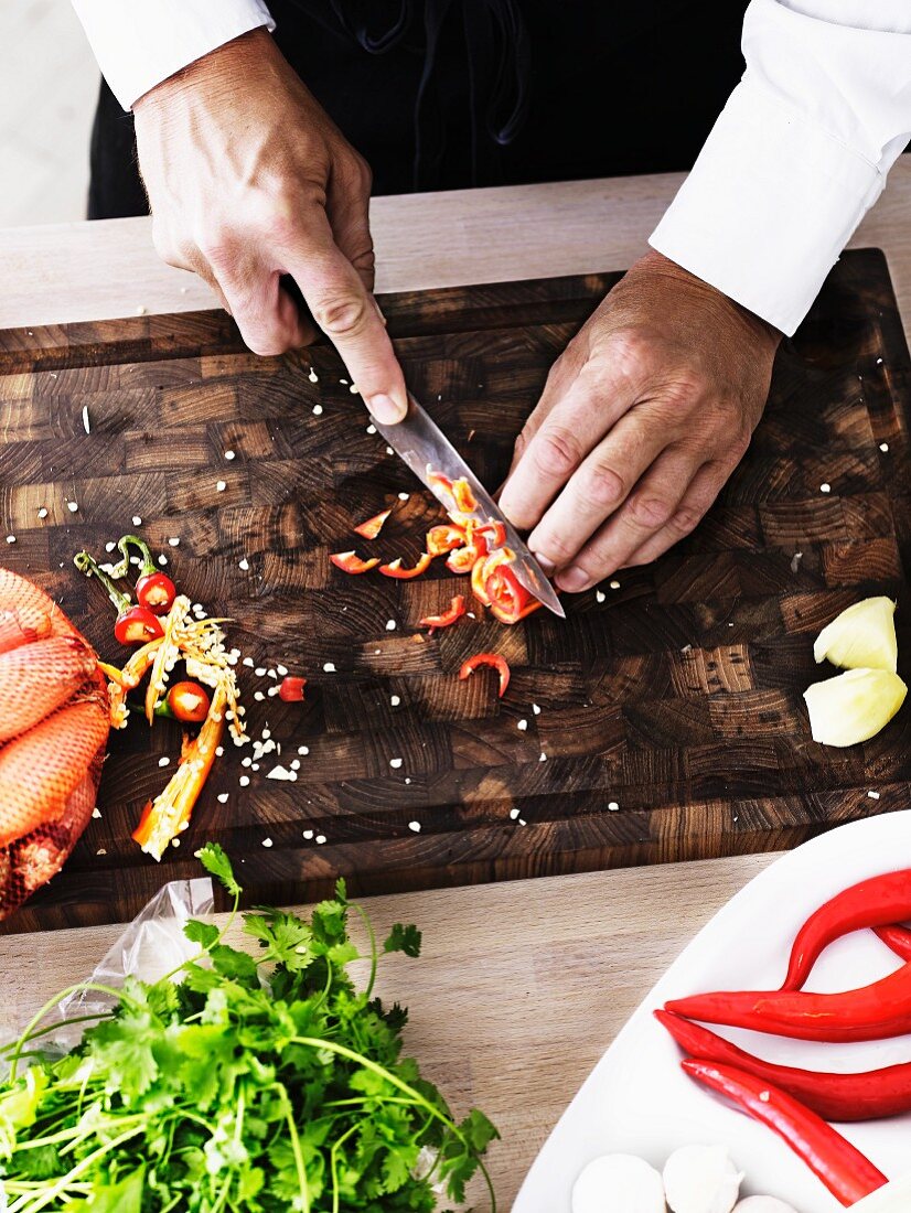 A man slicing chilli peppers into rings on a wooden board (seen from above)