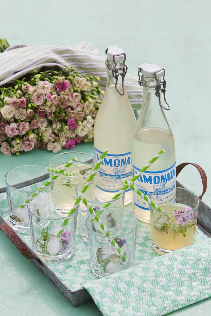 Glasses and vintage-style swing-top bottles of lemonade on simple metal tray in front of luxuriant bouquet of roses