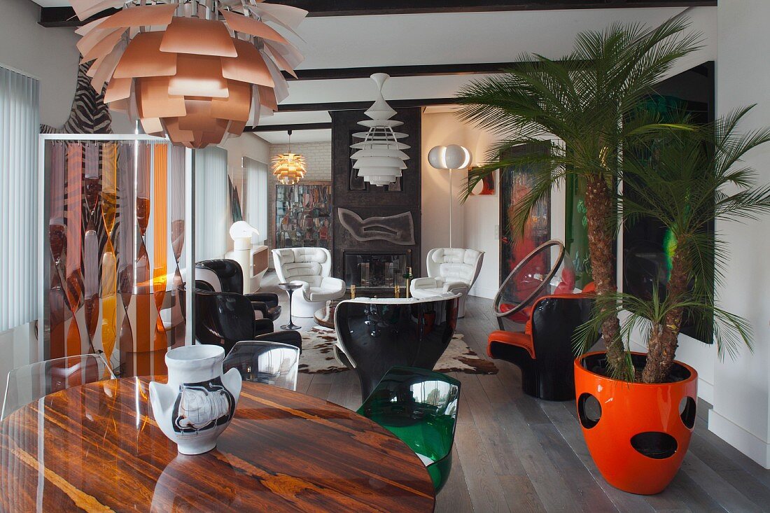 Round table below classic pendant lamp in front of lounge area with retro armchairs and palm in orange pot