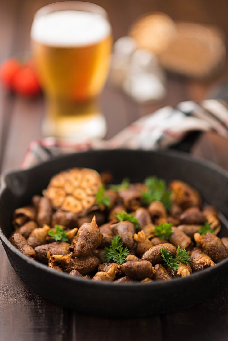 Fried chicken hearts with garlic and parsley in a pan with a glass of beer in the background