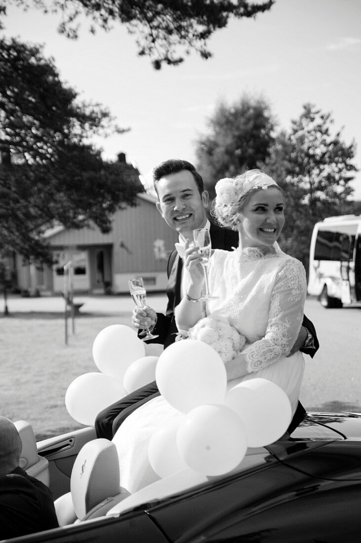 A bride and groom with champagne and balloons in a convertible (black-and-white shot)