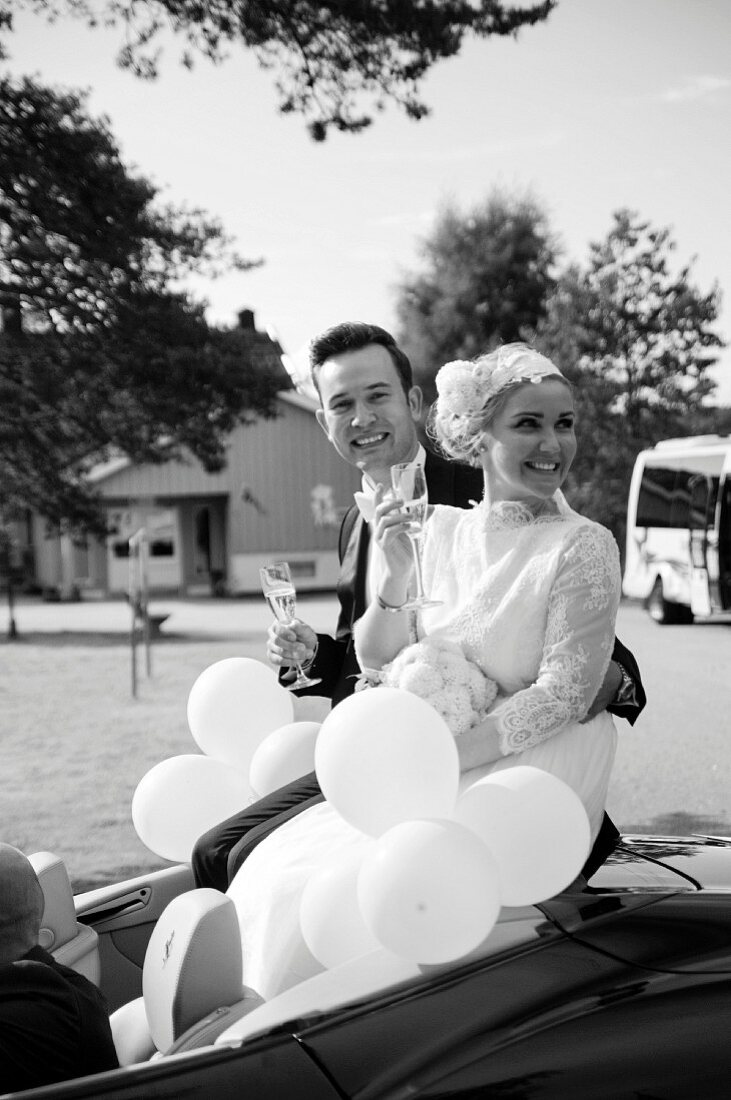 Hochzeitspaar mit Sekt und Ballons in offenem Cabrio (s/w-Bild)