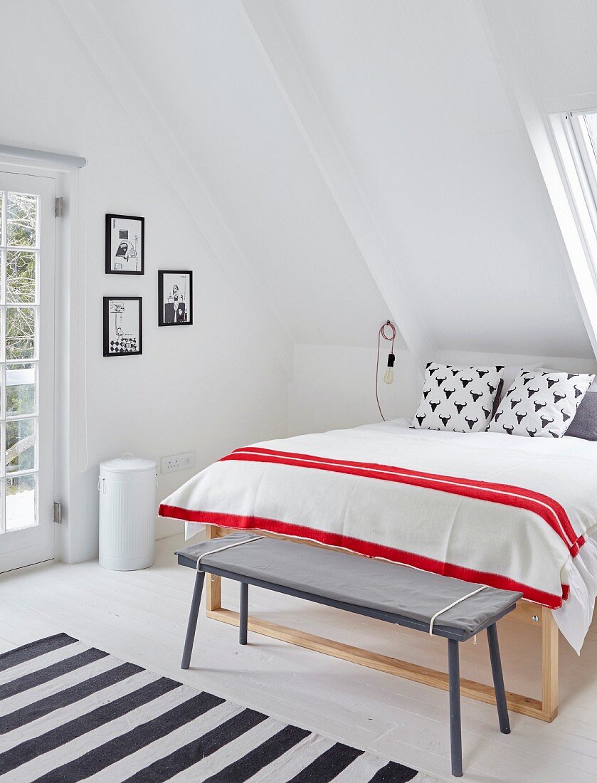 Simple bench at foot of double bed with red and white striped blanket below sloping ceiling