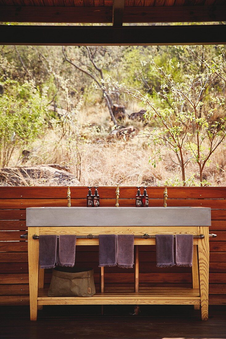 Concrete washstand on wooden base frame with towels hung on rail below panoramic window with view of natural landscape