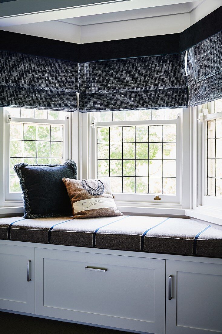 Cushioned window seat with cupboards below, lead glazed bay window and dark grey Roman blinds