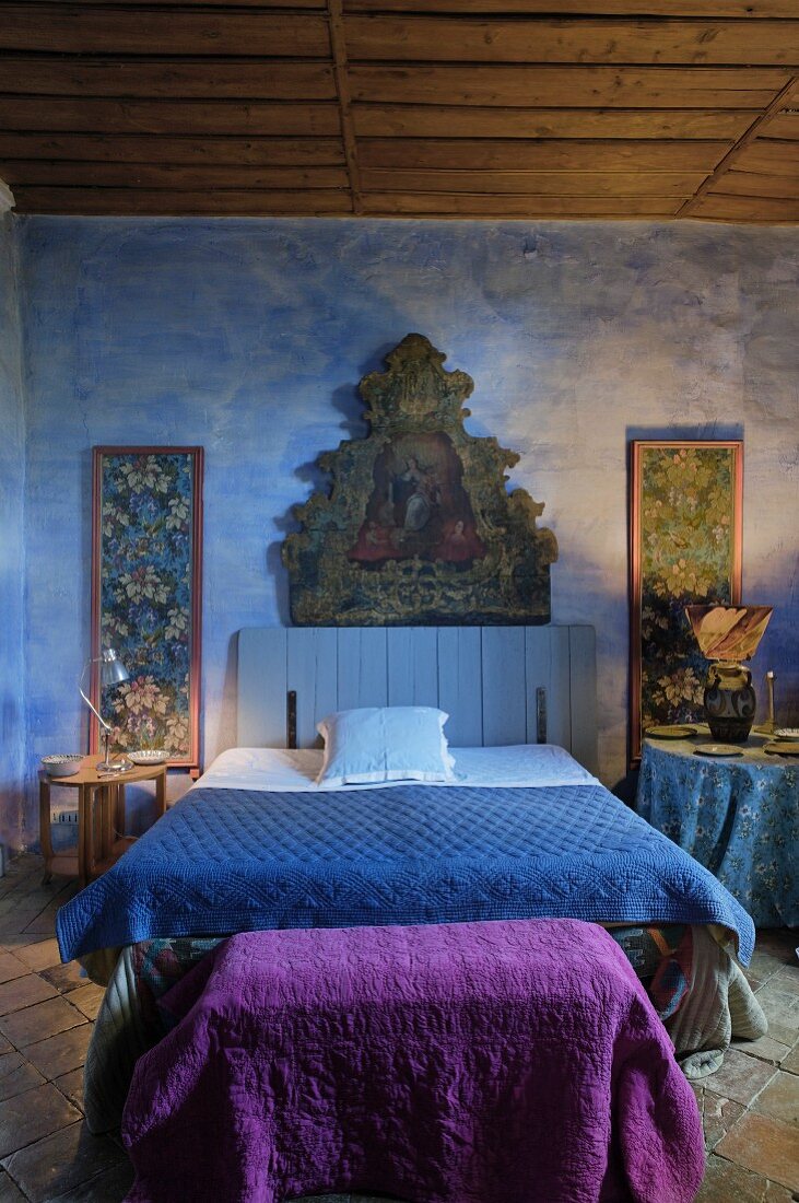 Ländliches Schlafzimmer mit religiösem Gemälde an blaugetönter Wand und Doppelbett mit blauer Tagesdecke, an Fussende violettes Plaid auf Truhe