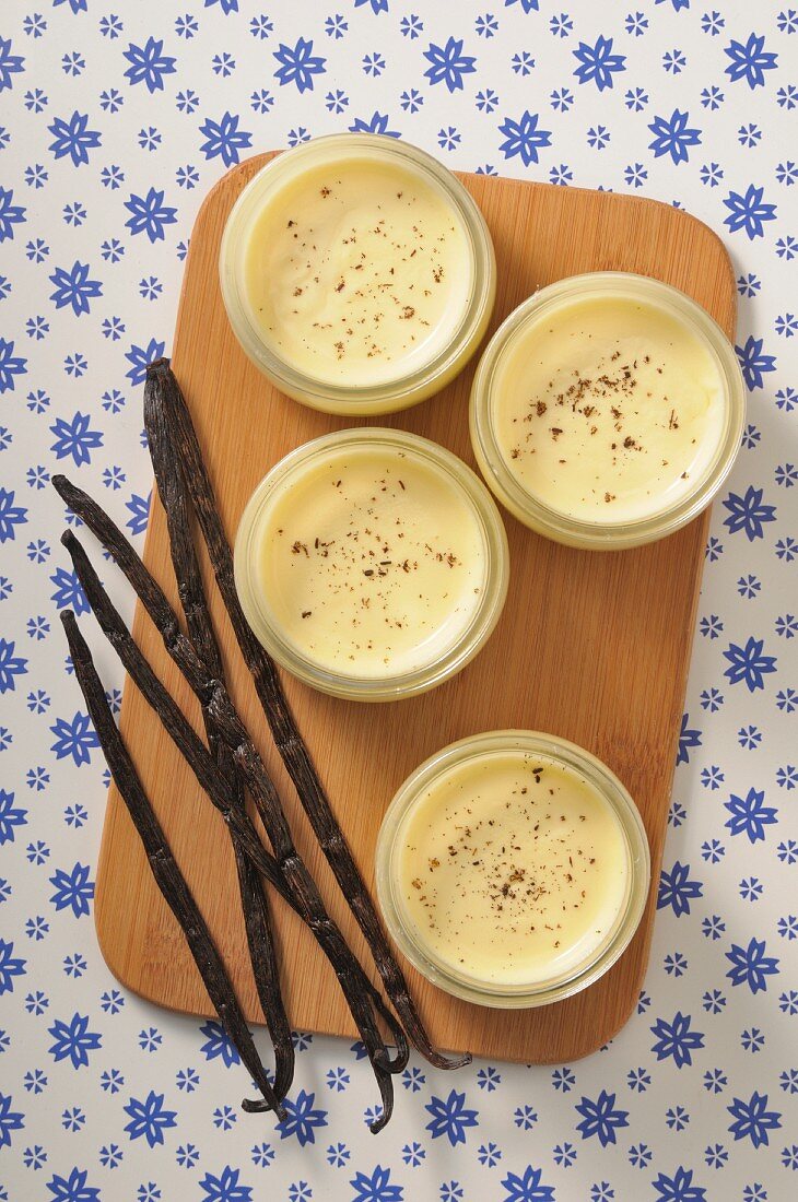 Four dishes of vanilla cream on a wooden board with vanilla pods