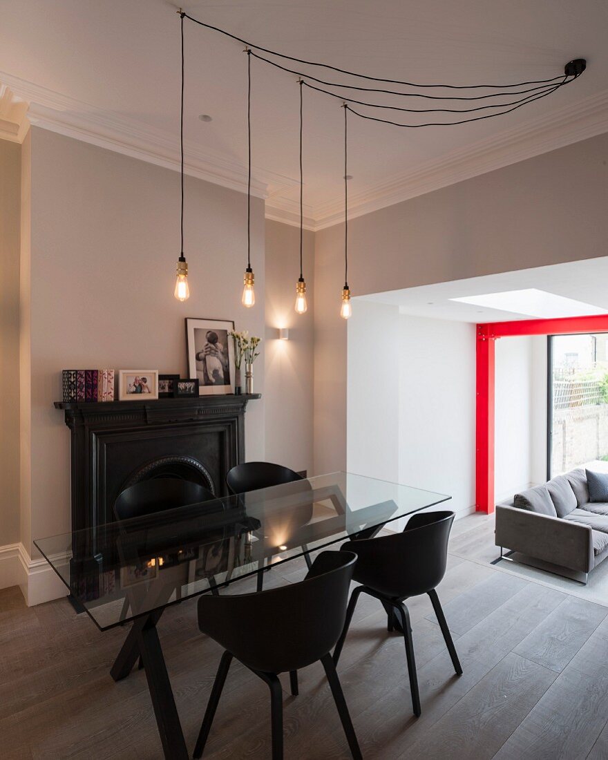 Light-bulb pendant lamp above glass table and black shell chairs in open-plan interior with red steel structure in background