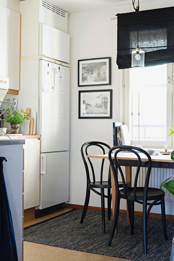 Black bistro chairs around wooden table in dining area in kitchen below window and next to white fitted fridge-freezer