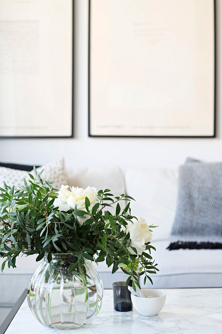 White peonies and leaves in glass vase on marble table in front of sofa