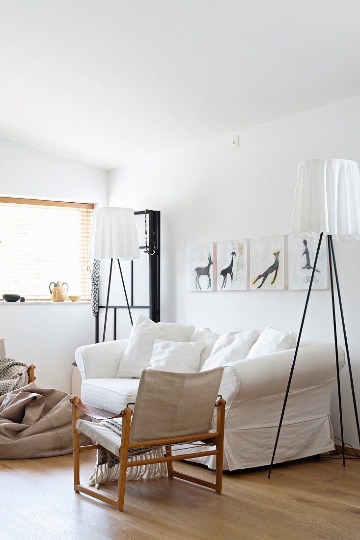 Designer standard lamp next to loose-covered sofa and armchair with wooden frame and linen seat and backrest in white living room