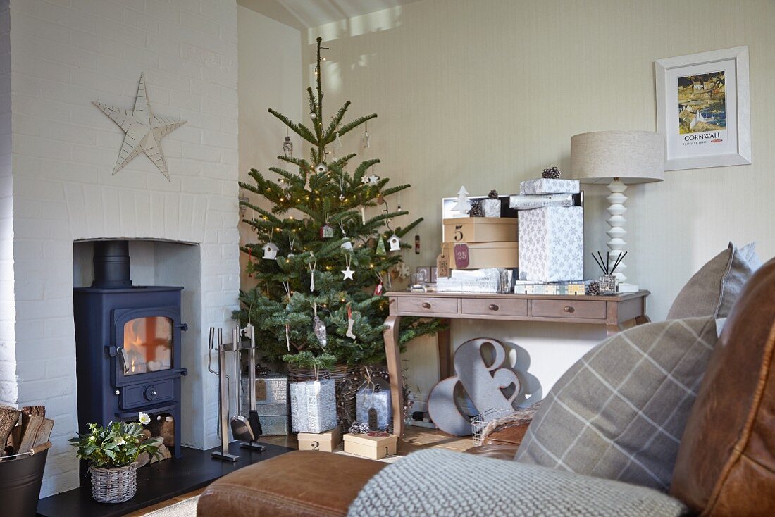 Decorated Christmas tree between wood-burning stove and gifts on console table in rustic living room