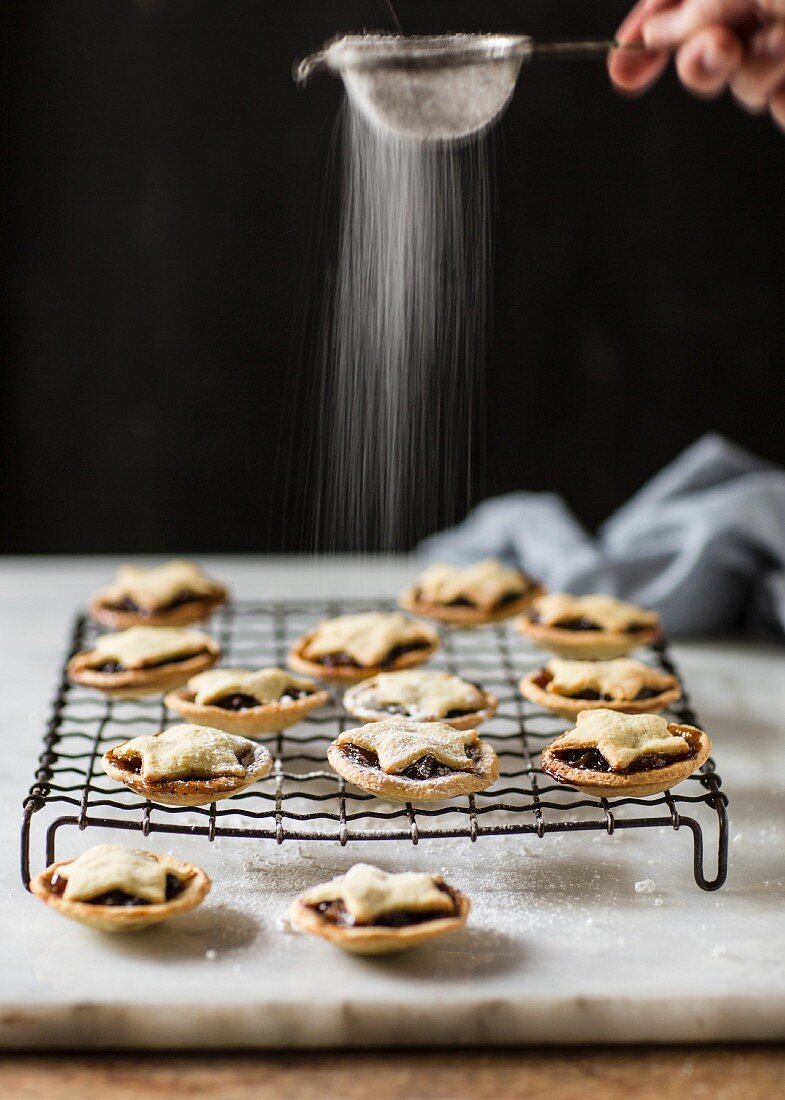 Mince pies on a cooling rack being dusted icing sugar