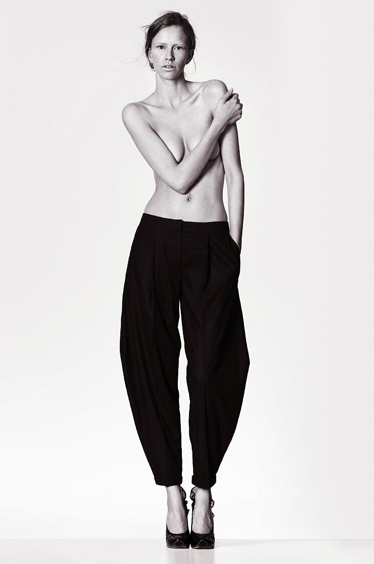 Young topless woman wearing baggy trousers covering breasts with one arm (black and white photo)