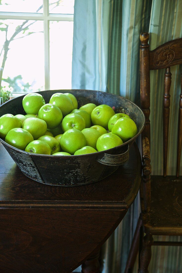 Green apples in a metal bowl on a wooden table in front of the window