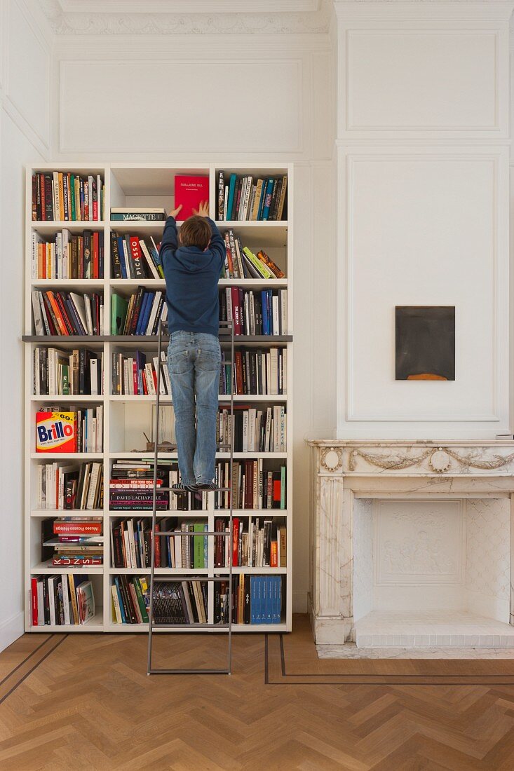 Boy climbing library ladder in front of bookcase next to fireplace with marble surround