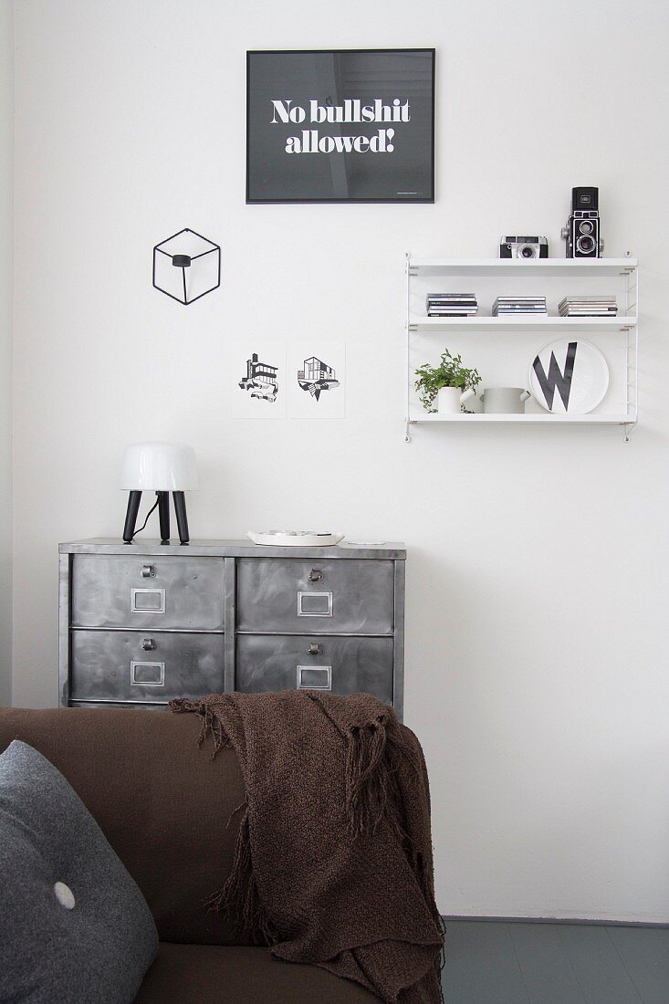 Brown sofa in front of metal chest of drawers below String shelves and decorations on wall