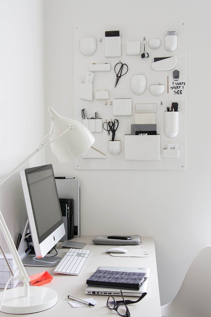 Monitor and white table lamp on white desk in corner below white, plastic retro organiser on wall