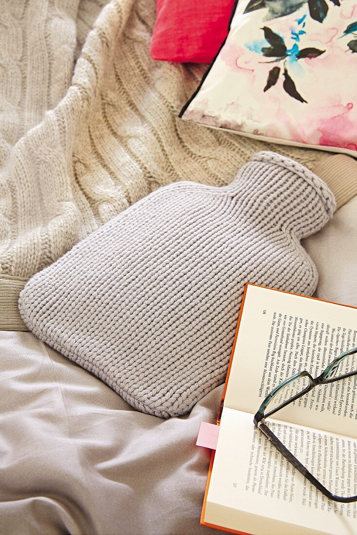 Hot-water bottle with pale grey knitted cover, blanket and book on bed